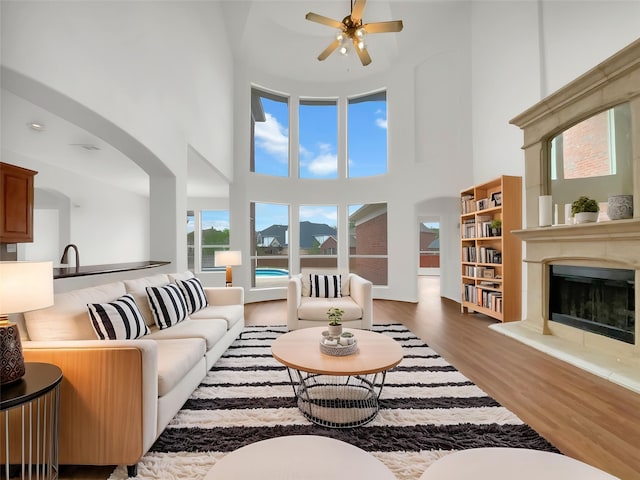 living area featuring ceiling fan, arched walkways, a towering ceiling, a fireplace with raised hearth, and wood finished floors