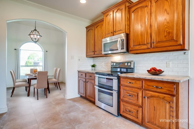 kitchen with brown cabinetry, backsplash, ornamental molding, arched walkways, and appliances with stainless steel finishes
