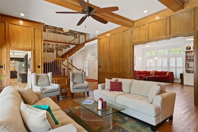 living room with beamed ceiling, a ceiling fan, wood finished floors, stairs, and wooden walls