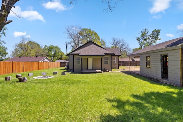back of property with a lawn, roof with shingles, a fenced backyard, and an outdoor fire pit