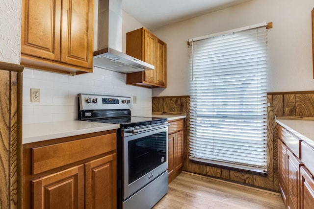kitchen featuring brown cabinetry, light wood-style flooring, wall chimney range hood, stainless steel electric range, and plenty of natural light
