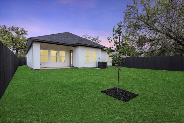 back of property at dusk with a shingled roof, a yard, a fenced backyard, and central AC unit