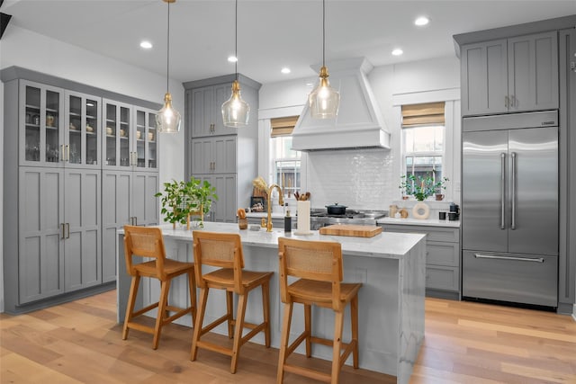 kitchen featuring light stone countertops, decorative backsplash, gray cabinets, built in refrigerator, and custom exhaust hood