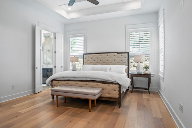 bedroom featuring ensuite bath, ceiling fan, baseboards, light wood-type flooring, and a tray ceiling