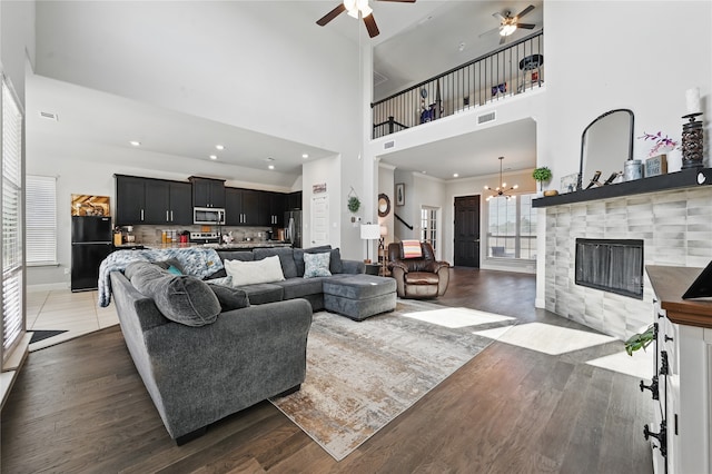 living room featuring visible vents, a towering ceiling, wood finished floors, a tiled fireplace, and ceiling fan with notable chandelier