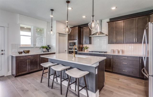 kitchen with wall chimney exhaust hood, a sink, appliances with stainless steel finishes, dark brown cabinetry, and light wood-style flooring