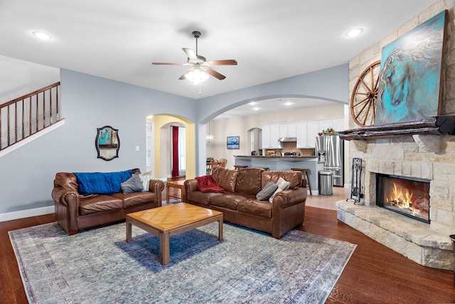 living room featuring a stone fireplace, ceiling fan, arched walkways, baseboards, and dark wood-style floors