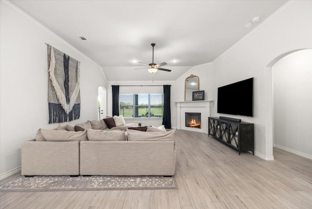 living room with arched walkways, ornamental molding, ceiling fan, light wood-style floors, and a lit fireplace