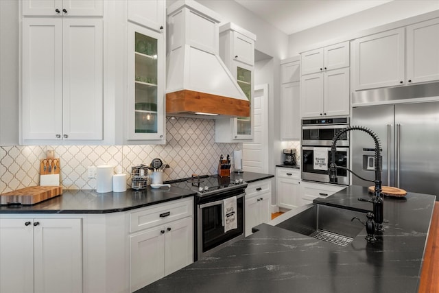 kitchen featuring custom exhaust hood, glass insert cabinets, white cabinets, and appliances with stainless steel finishes