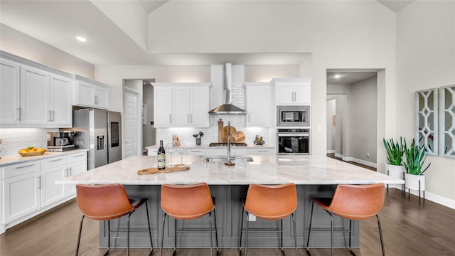 kitchen featuring wall chimney range hood, a kitchen bar, appliances with stainless steel finishes, tasteful backsplash, and white cabinets