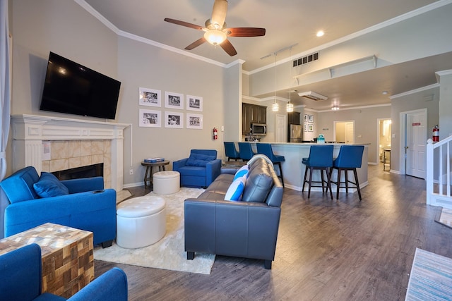 living area with ceiling fan, visible vents, a tiled fireplace, dark wood finished floors, and ornamental molding