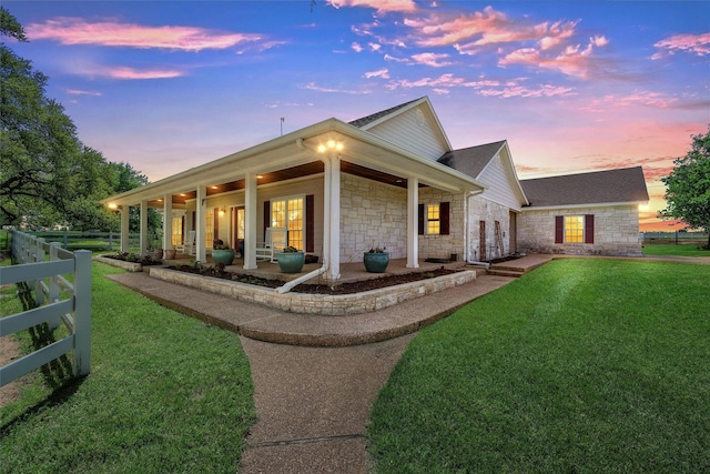 back of property at dusk featuring a lawn, fence, stone siding, and a shingled roof