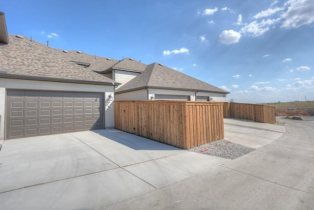 view of property exterior with concrete driveway, a shingled roof, an attached garage, and fence