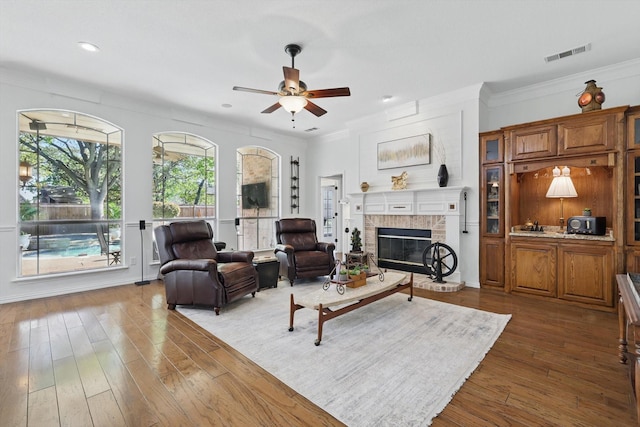 living area featuring crown molding, dark wood finished floors, a ceiling fan, visible vents, and a brick fireplace