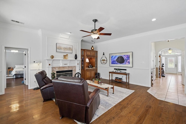 living area with crown molding, a ceiling fan, visible vents, a brick fireplace, and wood finished floors
