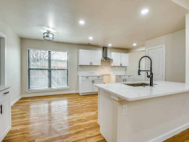 kitchen with wall chimney range hood, backsplash, white cabinets, light wood-style floors, and a sink