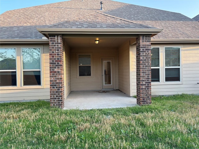 entrance to property featuring roof with shingles, a yard, a patio, and brick siding