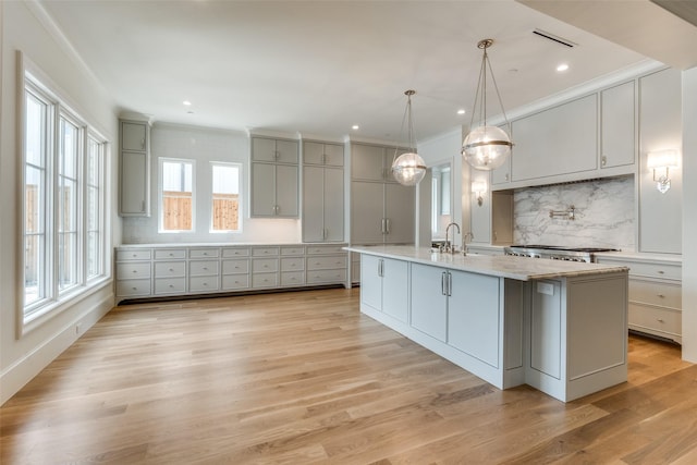 kitchen with gray cabinetry, decorative backsplash, an island with sink, light wood-style flooring, and crown molding