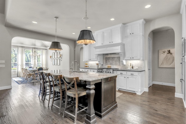 kitchen with custom exhaust hood, baseboards, an island with sink, arched walkways, and a sink