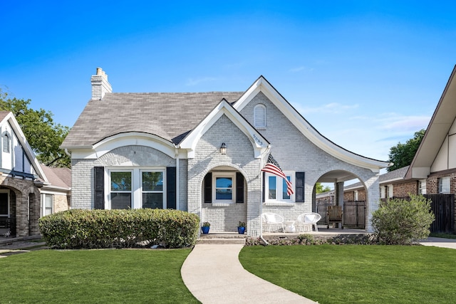 tudor house with a shingled roof, a chimney, fence, brick siding, and a front lawn