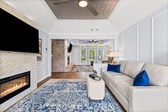 living room featuring a glass covered fireplace, a ceiling fan, visible vents, a raised ceiling, and wood finished floors