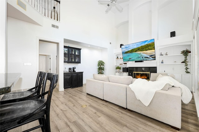 living room featuring visible vents, built in features, a glass covered fireplace, a high ceiling, and light wood-style flooring
