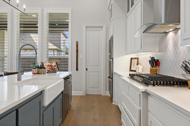 kitchen with wall chimney exhaust hood, light wood-style floors, decorative backsplash, light countertops, and appliances with stainless steel finishes