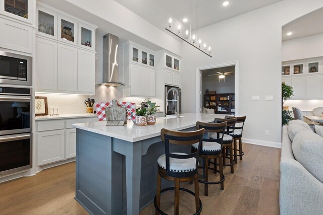 kitchen featuring an island with sink, backsplash, light countertops, wood finished floors, and wall chimney range hood