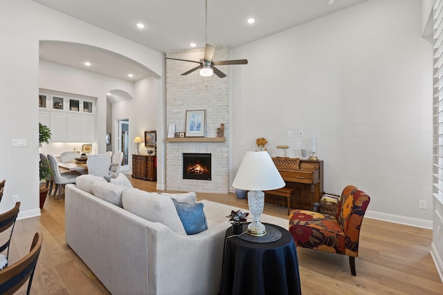 living room featuring light wood-type flooring, ceiling fan, arched walkways, a fireplace, and baseboards