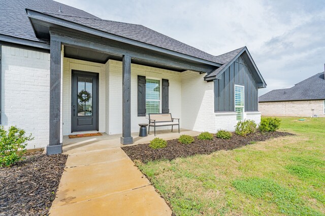 view of exterior entry featuring a yard, brick siding, a shingled roof, and covered porch