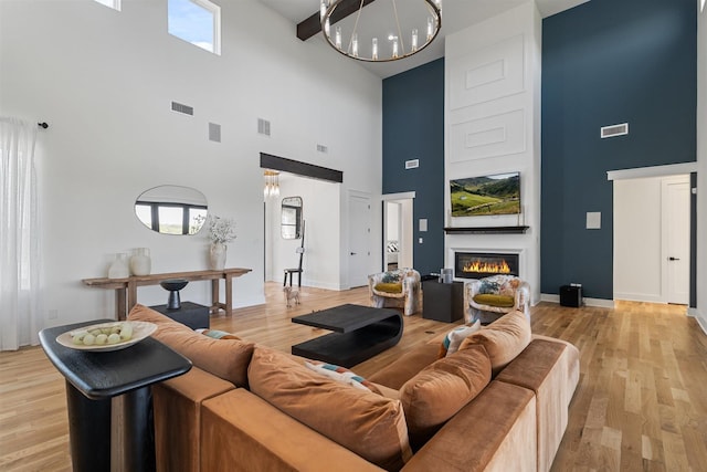 living room featuring a towering ceiling, an inviting chandelier, visible vents, a glass covered fireplace, and light wood-style flooring