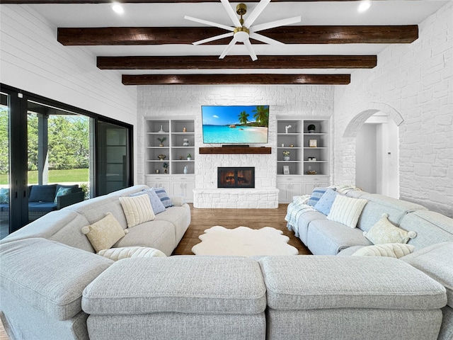 living area featuring a ceiling fan, a stone fireplace, arched walkways, beam ceiling, and wood finished floors