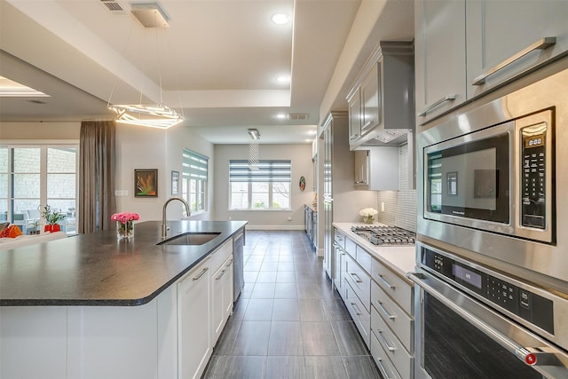 kitchen with a tray ceiling, dark countertops, a sink, stainless steel appliances, and a center island with sink