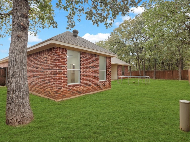 view of side of home with a trampoline, roof with shingles, brick siding, fence, and a lawn