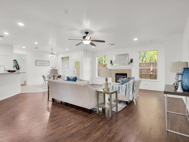 living area featuring dark wood-type flooring, ceiling fan with notable chandelier, a tiled fireplace, and recessed lighting