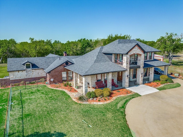 french country inspired facade featuring roof with shingles, a front yard, stone siding, a balcony, and a chimney