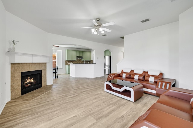 living area with a tiled fireplace, light wood-style flooring, a ceiling fan, visible vents, and arched walkways