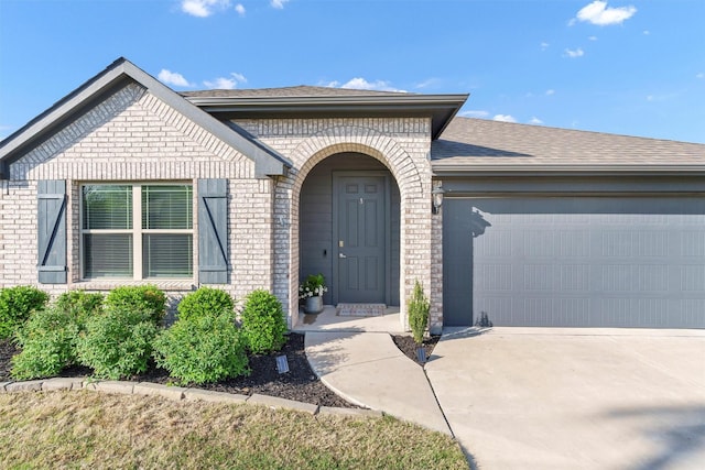 doorway to property featuring an attached garage, a shingled roof, driveway, and brick siding