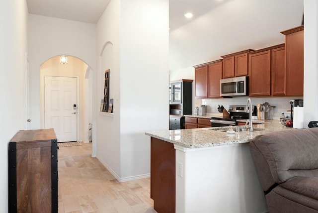 kitchen with arched walkways, a sink, brown cabinets, light stone counters, and appliances with stainless steel finishes