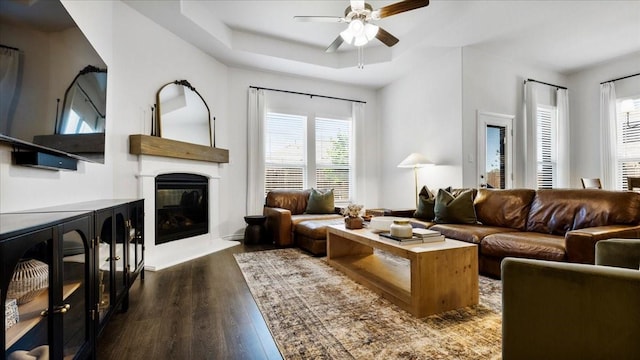living area featuring dark wood-style flooring, a ceiling fan, a glass covered fireplace, and a raised ceiling