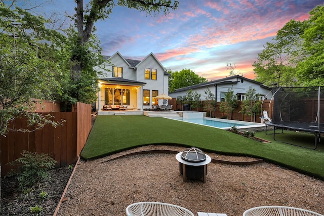 back of property at dusk with a fenced backyard, a trampoline, a fire pit, and a patio