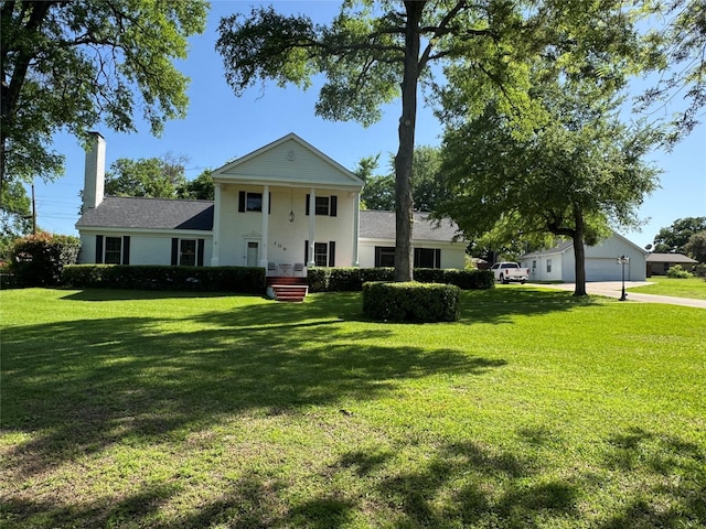 greek revival house with a chimney, a shingled roof, a detached garage, and a front yard