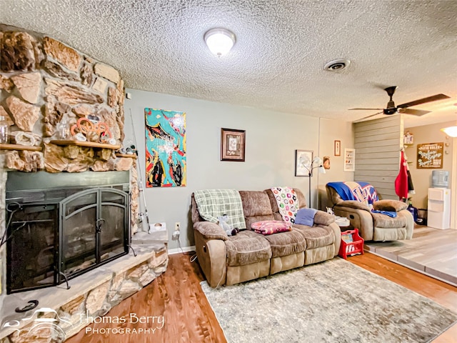 living room with wood finished floors, ceiling fan, a textured ceiling, and a stone fireplace