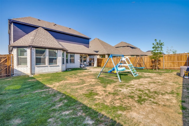 rear view of house with a shingled roof, a lawn, a fenced backyard, a playground, and brick siding