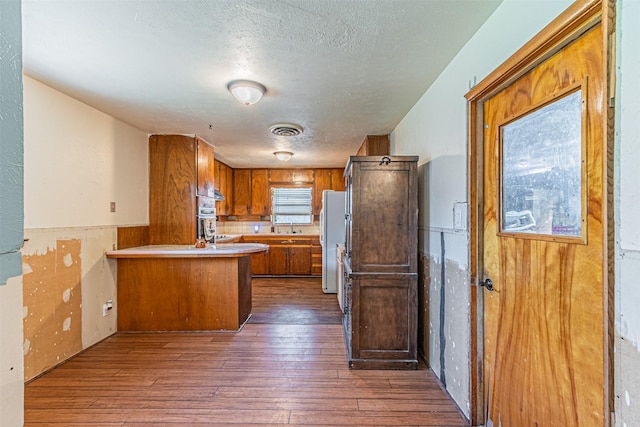 1206 John Reagan St, Benbrook, TX 76126 - kitchen with freestanding refrigerator, brown cabinetry, a peninsula, dark wood finished floors, and a textured ceiling