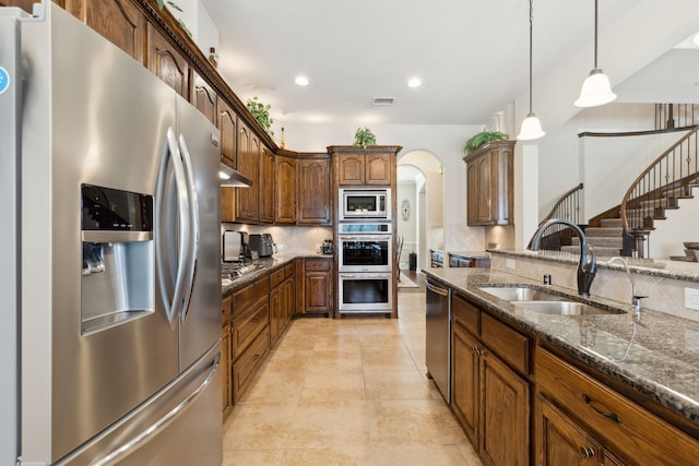 kitchen featuring arched walkways, dark stone countertops, appliances with stainless steel finishes, a sink, and backsplash