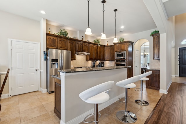 kitchen with light tile patterned floors, arched walkways, a kitchen island with sink, stainless steel appliances, and backsplash