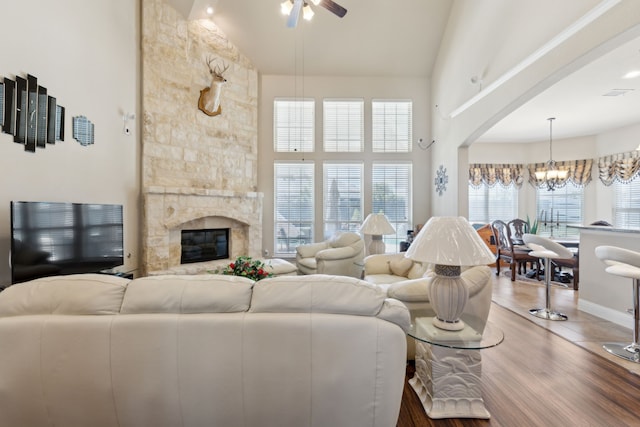 living area with ceiling fan with notable chandelier, high vaulted ceiling, arched walkways, a stone fireplace, and wood finished floors