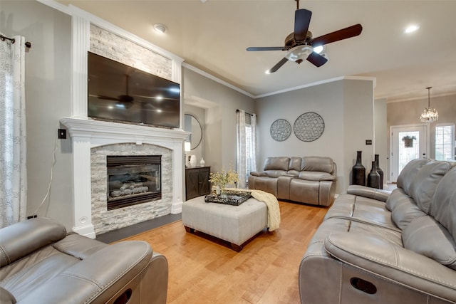 living room with light wood-style floors, ceiling fan with notable chandelier, crown molding, and a stone fireplace
