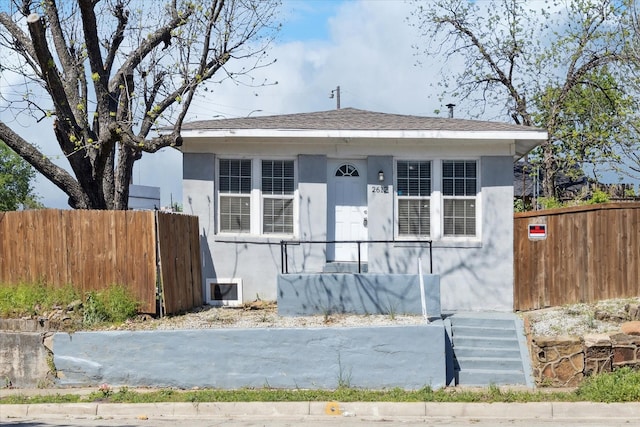 bungalow-style house with a shingled roof, stucco siding, and fence